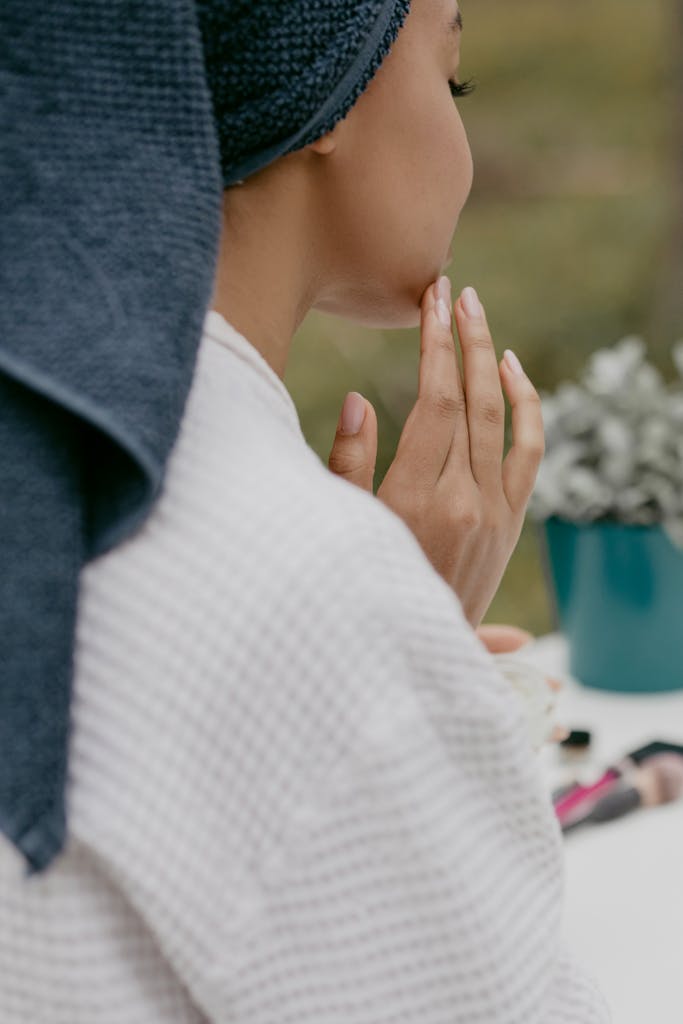 Back view of a woman applying facial cream, wearing a bath towel wrap, promoting healthy skin care.