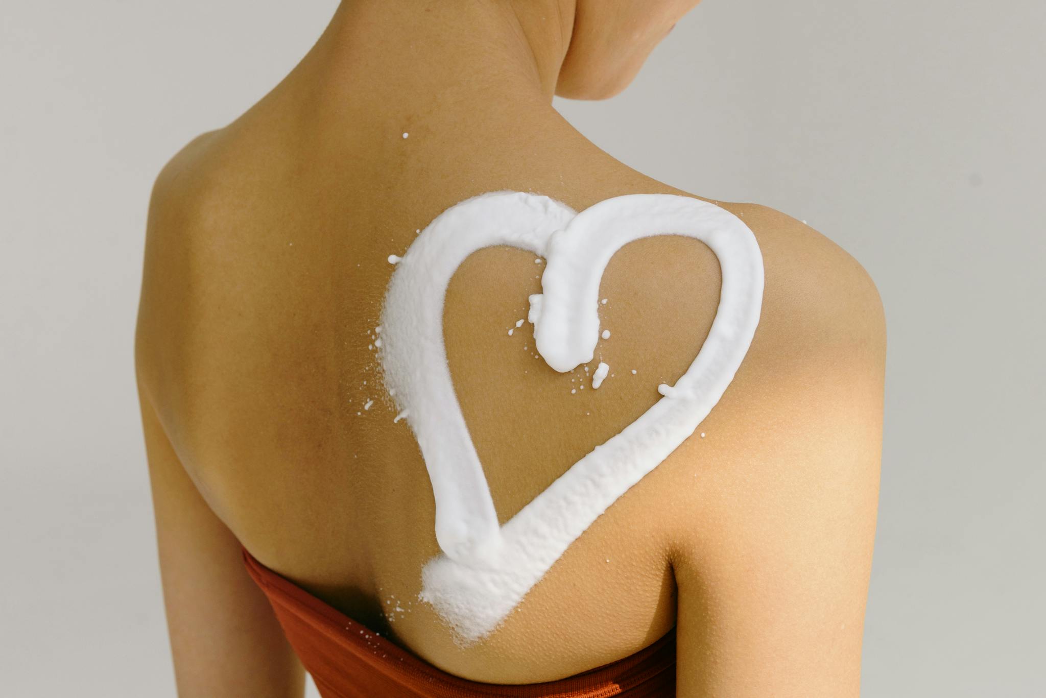 Rear view of a woman's shoulder with a heart shape drawn in cream, symbolizing love and skincare.