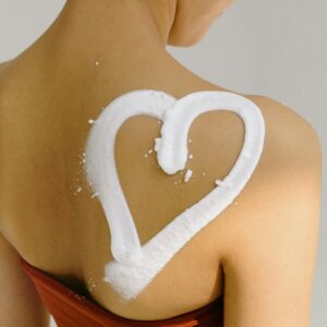 Rear view of a woman's shoulder with a heart shape drawn in cream, symbolizing love and skincare.